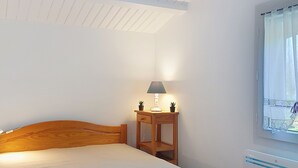 A neatly made bed with a blue bedspread, a wooden headboard, and a bedside table with a lamp and two potted plants. - La Coti 3 by Interhome (Ile d'Oléron)