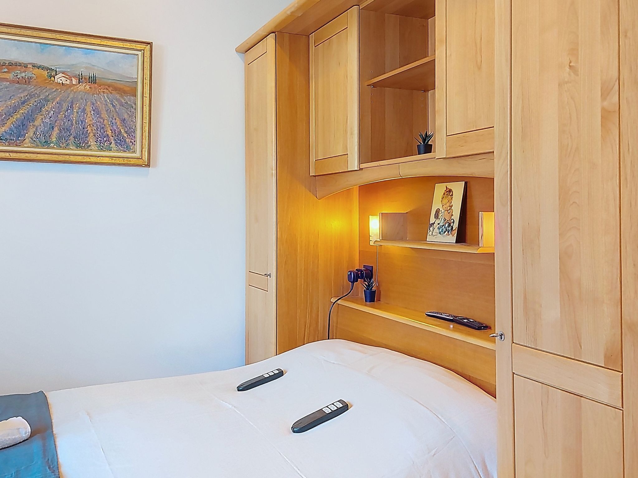 A bedroom with a bed, wooden wardrobe, a painting, a bedside lamp, and a small potted plant.
