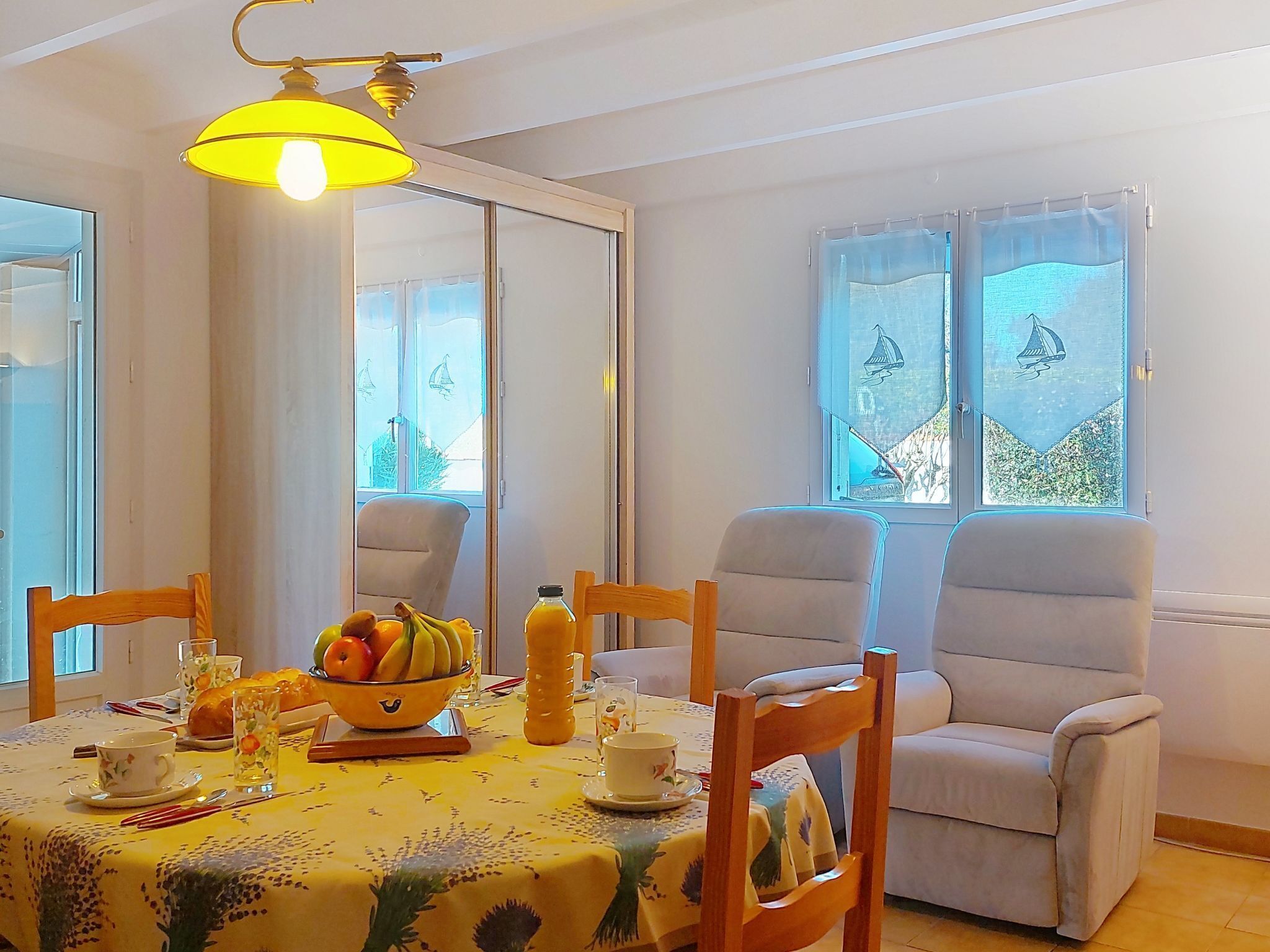 A dining area with a table set for two, featuring a bowl of fruit, a bottle of orange juice, and a floral tablecloth.