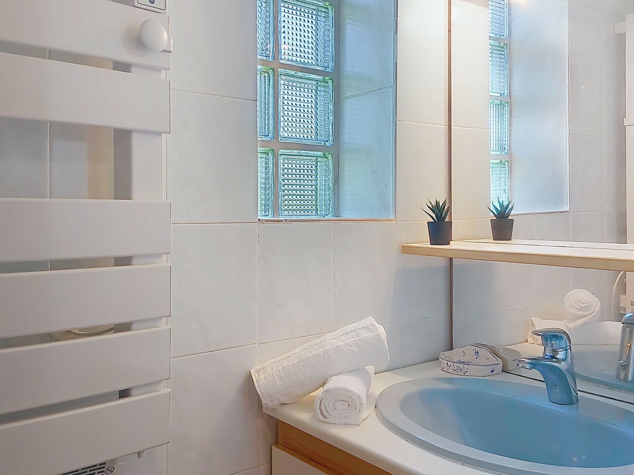 Un baño con paredes de azulejos blancos, un lavabo azul, un espejo, un perchero para toallas y una pequeña planta en maceta.