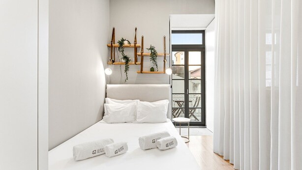 Une chambre moderne avec un lit, du linge de lit blanc, une table de chevet avec une lampe et une vue sur le balcon extérieur.