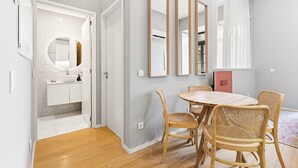 Un appartement moderne doté d’un coin repas avec une table ronde et des chaises en osier, d’une cuisine avec des meubles blancs et un dosseret carrelé, et d’un espace de vie avec une grande fenêtre et une pancarte indiquant « PRÊT POUR LES INVITÉS ».