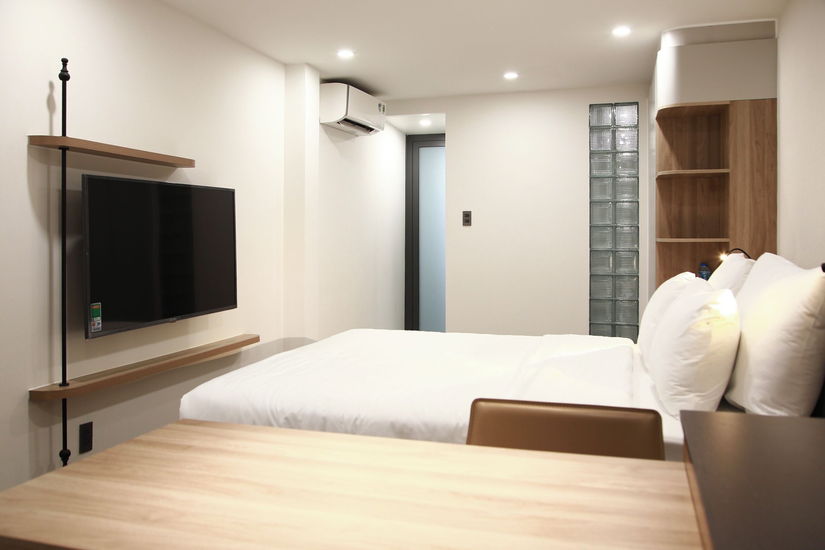 Quarto de hotel com cama, televisão fixada na parede, mesa de madeira e guarda-roupa com prateleiras.