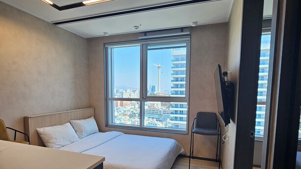 A modern bedroom with a large bed, a desk, and a chair. There is a window with a city view. - 용두산공원 뷰 및 바다뷰, 지하철역 2분거리!! 숙소 바로 앞 먹자골목, 국제시장 6분거리!! (Busan)