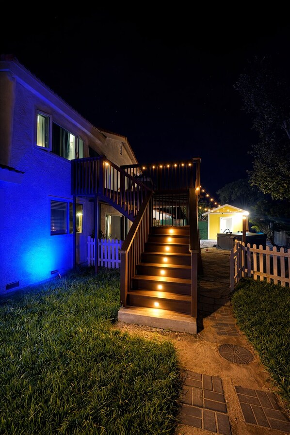 A house with a wooden staircase leading to a porch, surrounded by a white picket fence and a well-lit pathway.