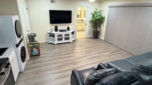 A living room with a flat-screen TV, a ceiling fan, a washing machine, and a dining area with a table and chairs.