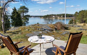 Eine Holzterrasse mit zwei Stühlen und einem Tisch, mit Blick auf einen See mit steiniger Uferlinie und Bäumen.