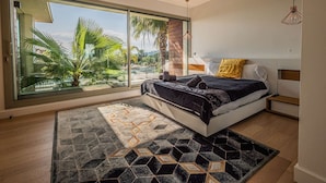 Un dormitorio moderno con un ventanal grande, una cama con colcha estampada, una alfombra geométrica y una lámpara colgante.