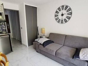 A modern living room with a grey sofa, a large wall clock, and a small side table with a lamp. - JARDINS D'YS Studio for 2 people with air conditioning (LES ISSAMBRES)