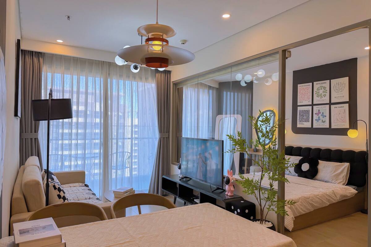 Una habitación de hotel moderna con una cama grande, un escritorio con televisor, un comedor y un amplio ventanal con cortinas.
