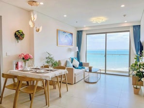 A modern living room with a dining area, a sofa, a coffee table, and a view of the sea. - Coral Condotel The Song - Vung Tau (Vũng Tàu)