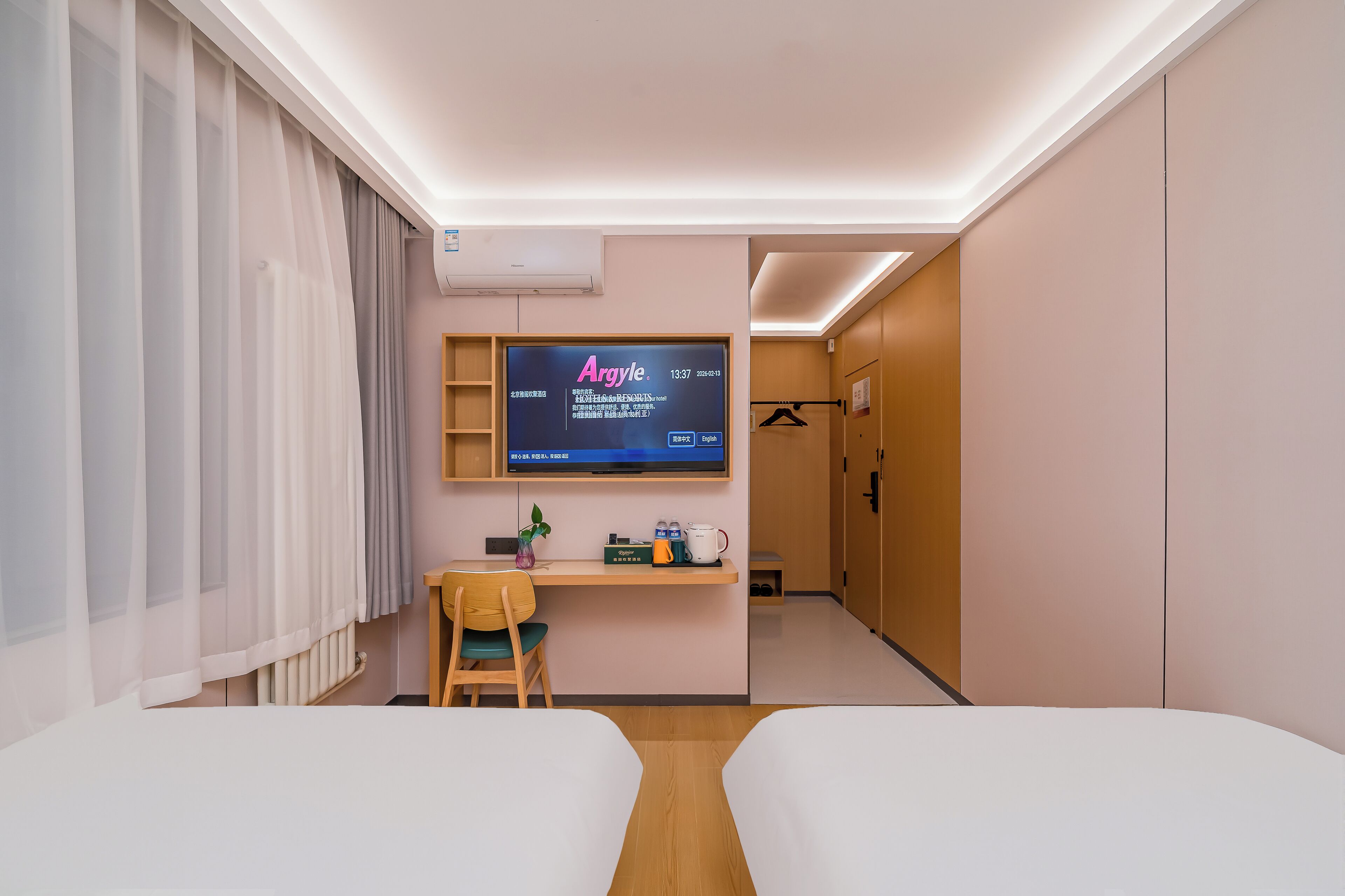 A hotel room with two beds, a desk with a chair, a television mounted on the wall, and a small plant on the desk.