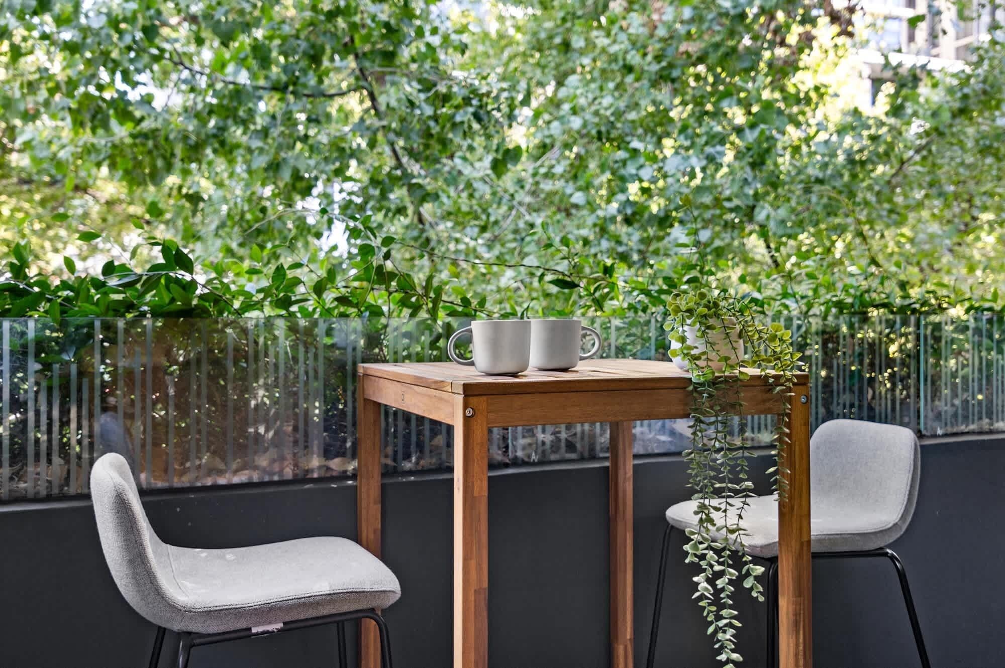 Une table en bois, sur laquelle reposent deux tasses blanches et une plante en pot, est encadrée par deux chaises grises, le tout se détachant sur un fond de feuillage verdoyant et luxuriant.