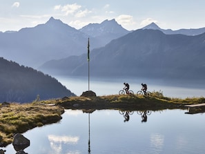 Un lago de montaña con dos ciclistas en un sendero, rodeado de montañas y un asta de banderín.