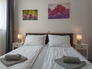 Un lit double avec du linge de lit blanc, deux oreillers et deux tables de chevet avec des lampes. Deux tableaux sont accrochés au mur au-dessus du lit.
