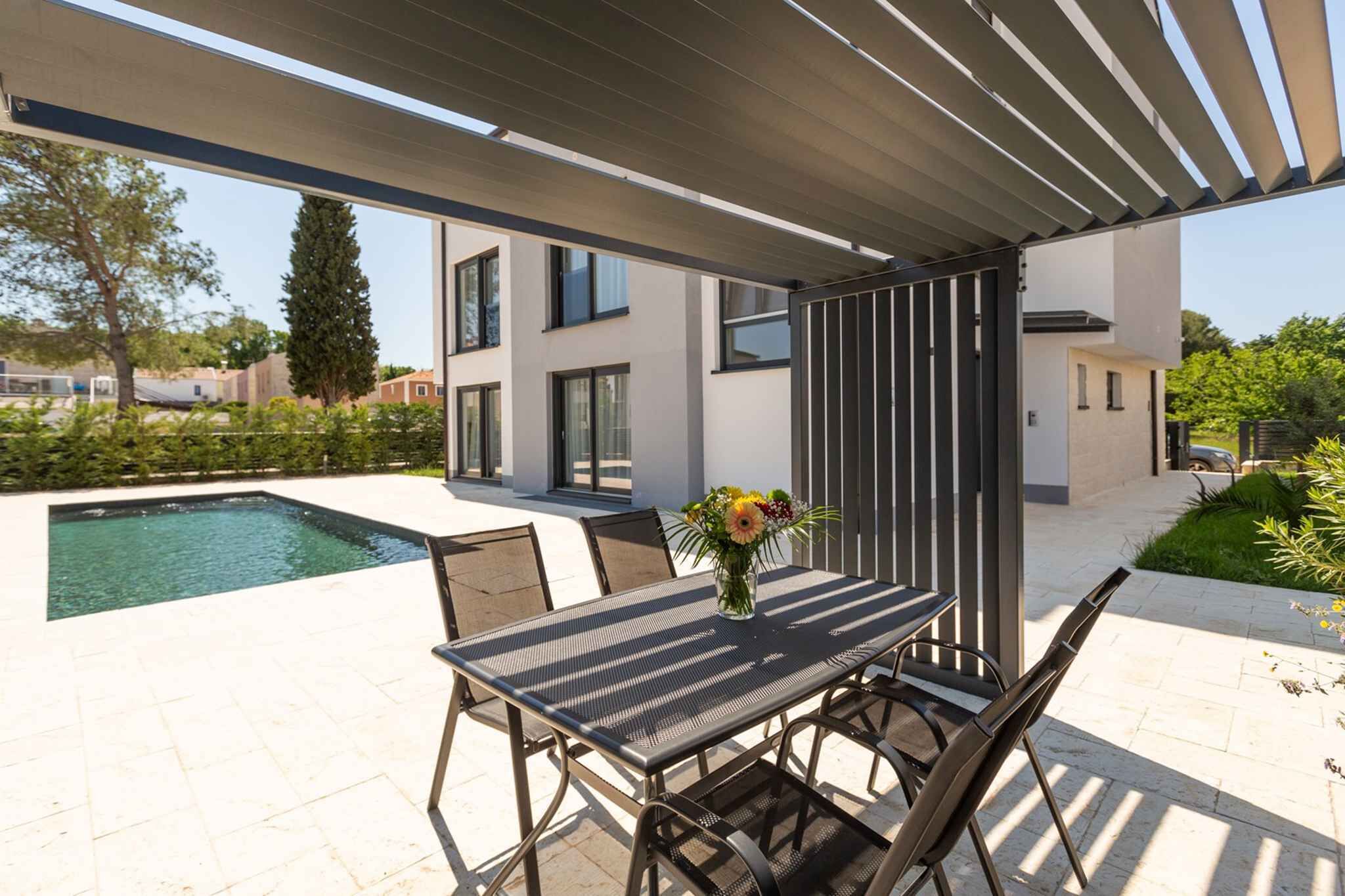 Um pátio moderno ao ar livre com piscina, mesa de jantar e cadeiras, e uma pérgula.