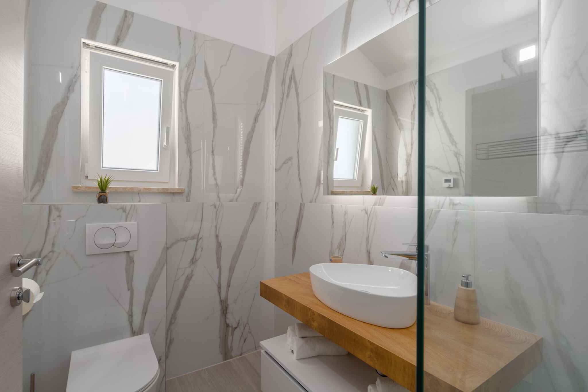 Un baño con paredes de mármol, un lavamanos blanco, tocador de madera, cabina de ducha de vidrio y un inodoro.