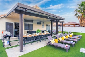 A covered outdoor area with a barbecue, dining table, and lounge chairs. - Spacious Group Vacation: Pool, Spa, Golf, Billiard (North Las Vegas)