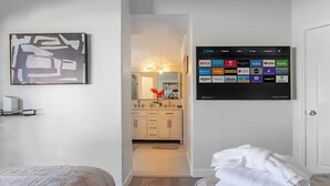 Hotellihuone, jossa on sänky, yöpöytä, seinälle asennettu televisio, jossa näkyy sovellusten kuvakkeet, sekä kylpyhuone, jossa on pesuallas ja peili.