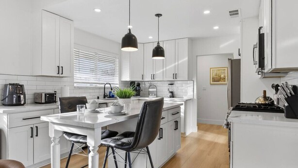 Une cuisine moderne avec des meubles blancs, une table à manger avec des chaises et des luminaires suspendus.