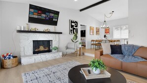 Un salon moderne doté d’une cheminée, d’un téléviseur à écran plat fixé au-dessus, d’un coin repas avec une table et des chaises, et d’une grande fenêtre avec des stores.