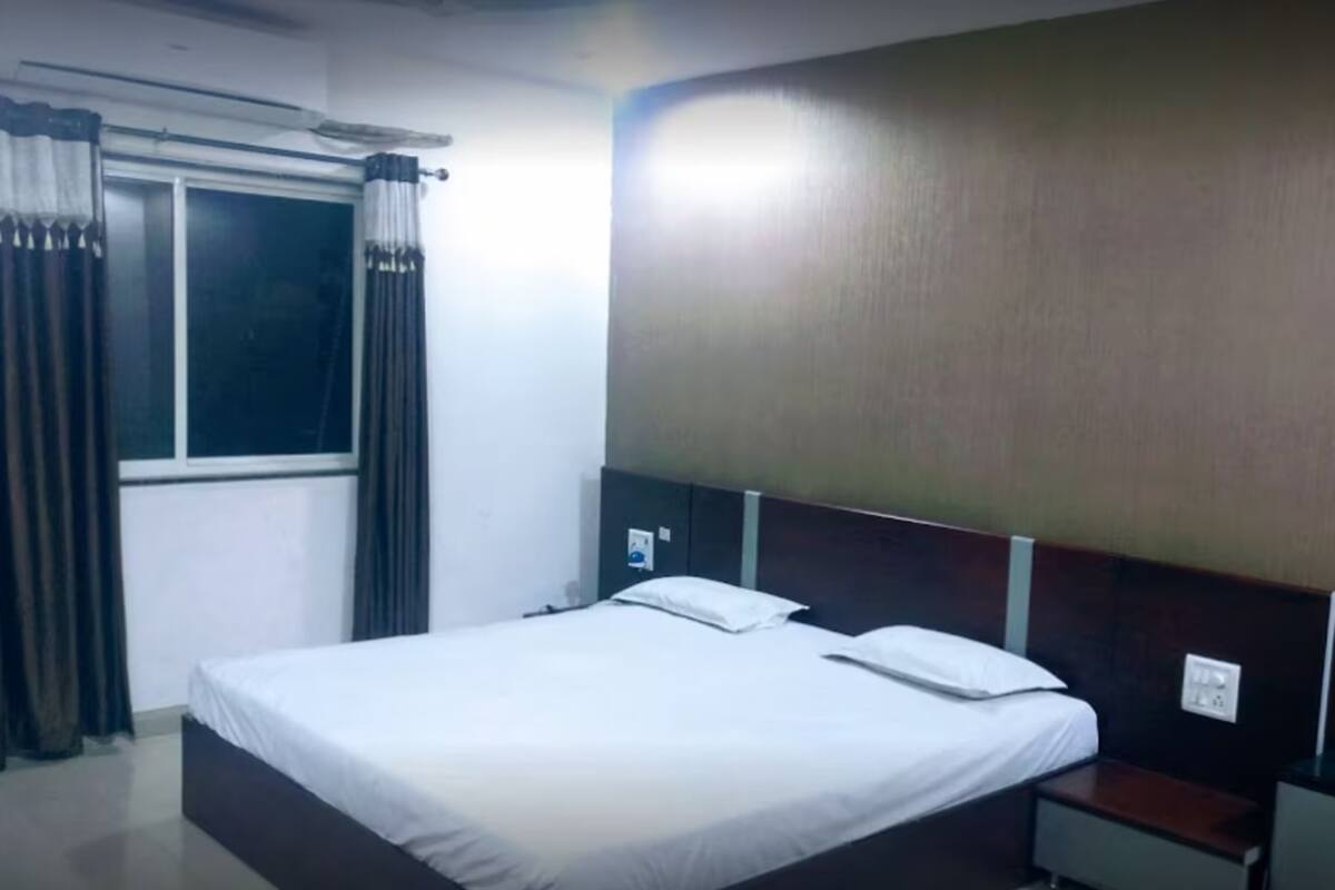 A hotel room with a single bed, a nightstand, a window with curtains, and a wall-mounted air conditioning unit.