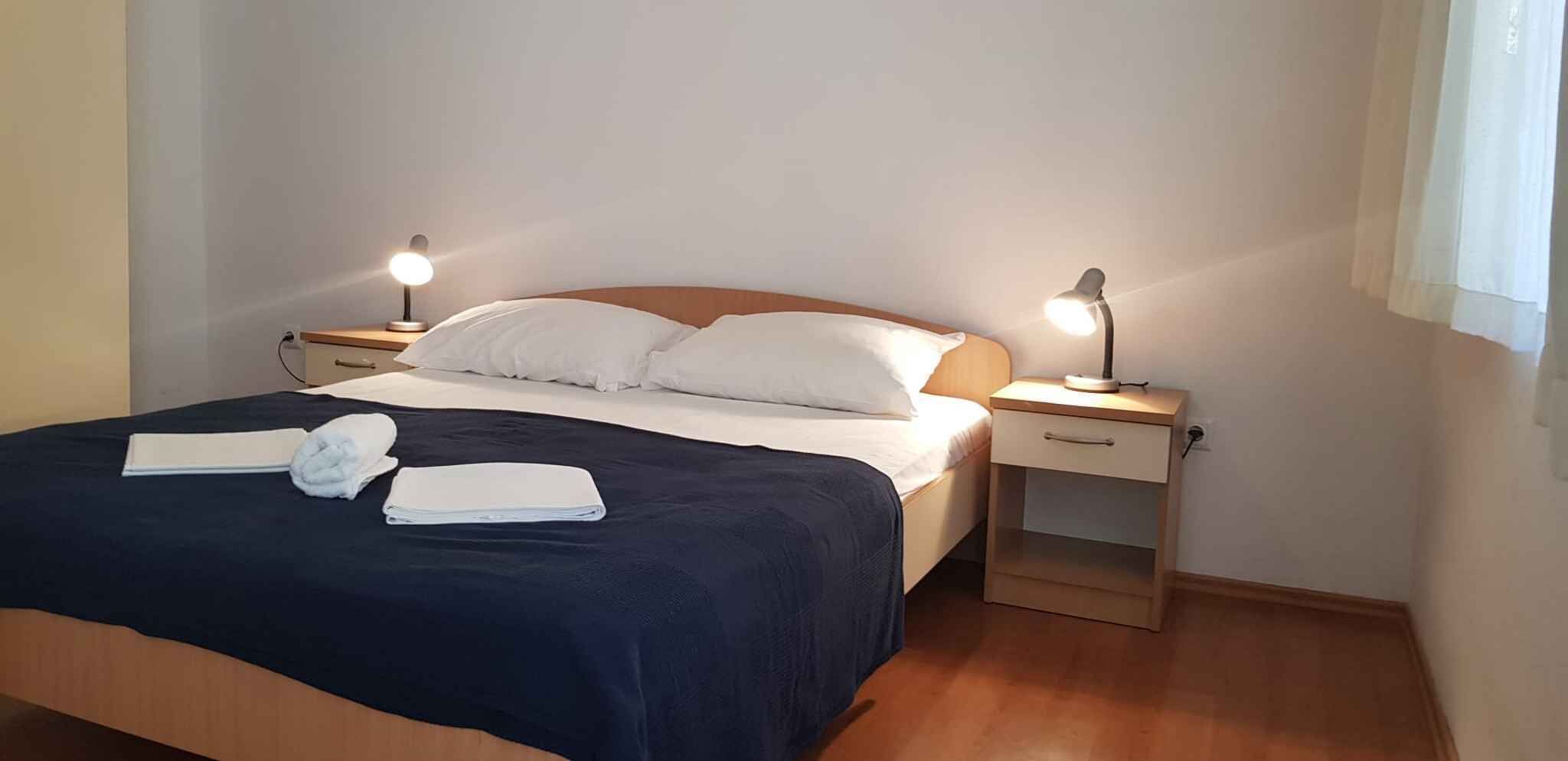 Una cama bien hecha con sábanas blancas y una colcha azul oscuro, dos mesitas de noche con lámparas, y una ventana con cortinas.
