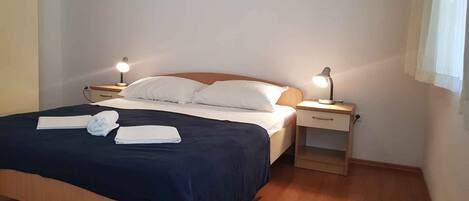 Una cama bien hecha con sábanas blancas y una colcha azul oscuro, dos mesitas de noche con lámparas, y una ventana con cortinas.