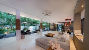 Un vaste espace de vie comprenant un grand canapé, une table basse et un ventilateur de plafond. On aperçoit à l’arrière-plan des parasols et une piscine.