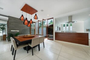 A modern kitchen with a wooden island, a dining area with a large table and chairs, and pendant lighting. - The Muse Luxury Eco-Living (Tabanan)