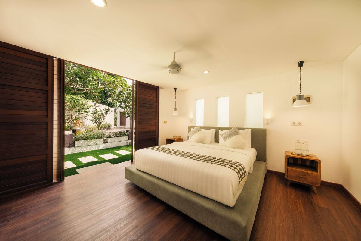 A modern bedroom with a large bed, wooden flooring, and a view of a garden through an open door.