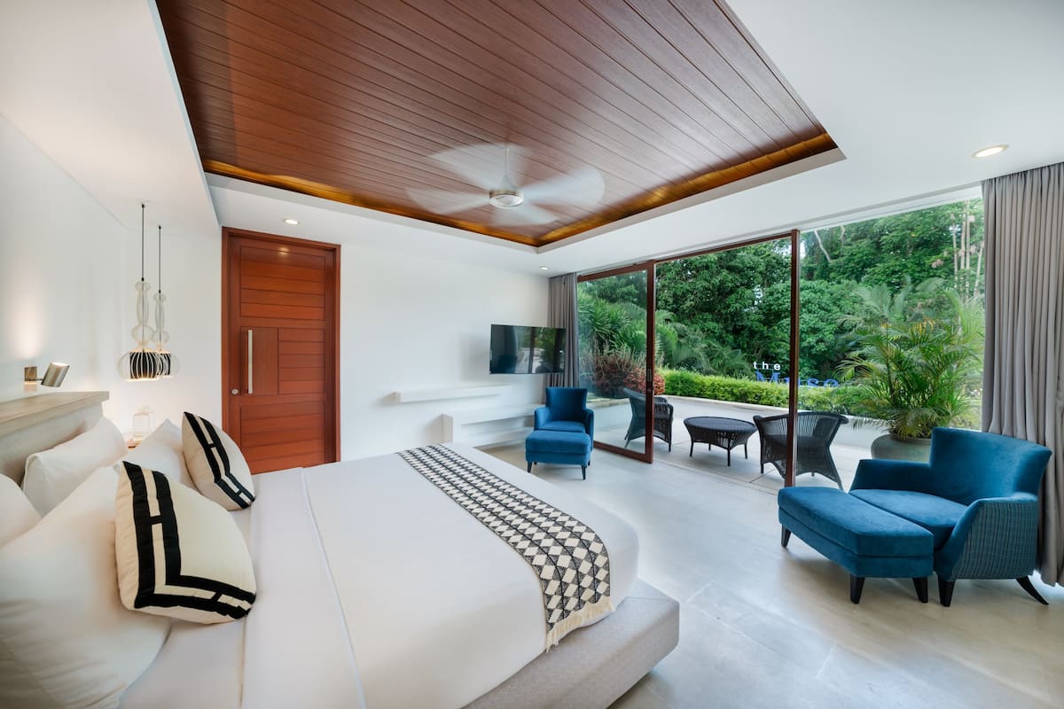A modern bedroom with a large bed, a ceiling fan, and a TV. There is a balcony with outdoor seating and a view of greenery.