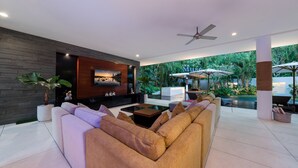 Un salon moderne avec un grand canapé, une table basse et un téléviseur fixé à un mur sombre. On aperçoit une piscine à l’arrière-plan.
