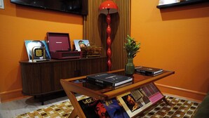 A room with a wooden coffee table displaying books and magazines, a red lamp, a small plant, and a wooden cabinet with a television above it. - The Tangerine Retreat - Retro Dallas Downtown Vibe (Dallas)