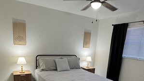 Une chambre à coucher comprenant un lit, deux tables de chevet, un ventilateur de plafond et une fenêtre avec des stores.