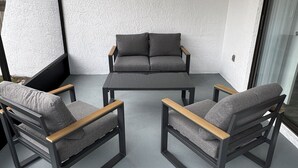 Un balcon avec un canapé gris et deux fauteuils, une petite table et une porte vitrée.