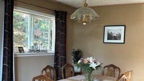 Een eetruimte met een tafel voor zes personen, omringd door houten stoelen, een kroonluchter, een ingelijst schilderij aan de muur en een raam met uitzicht op bomen.