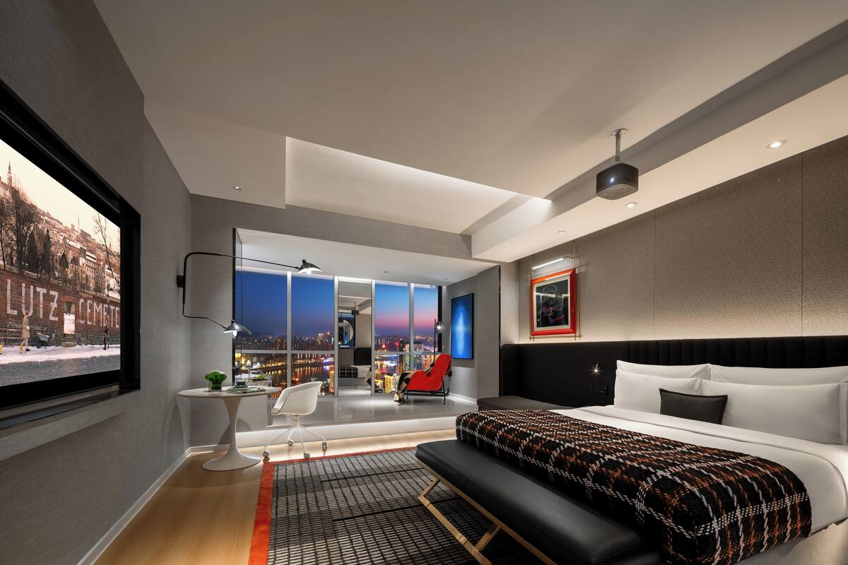 Une chambre d’hôtel moderne, dotée d’un grand lit, d’un bureau, d’une chaise et offrant une vue sur le paysage urbain.