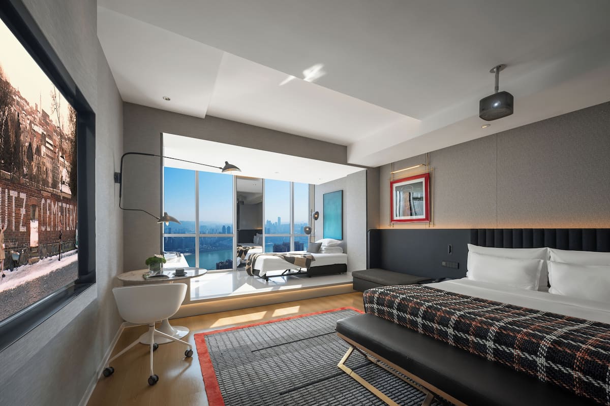 Habitación de hotel moderna con una ventana grande, una cama, un escritorio y vistas a un paisaje urbano.