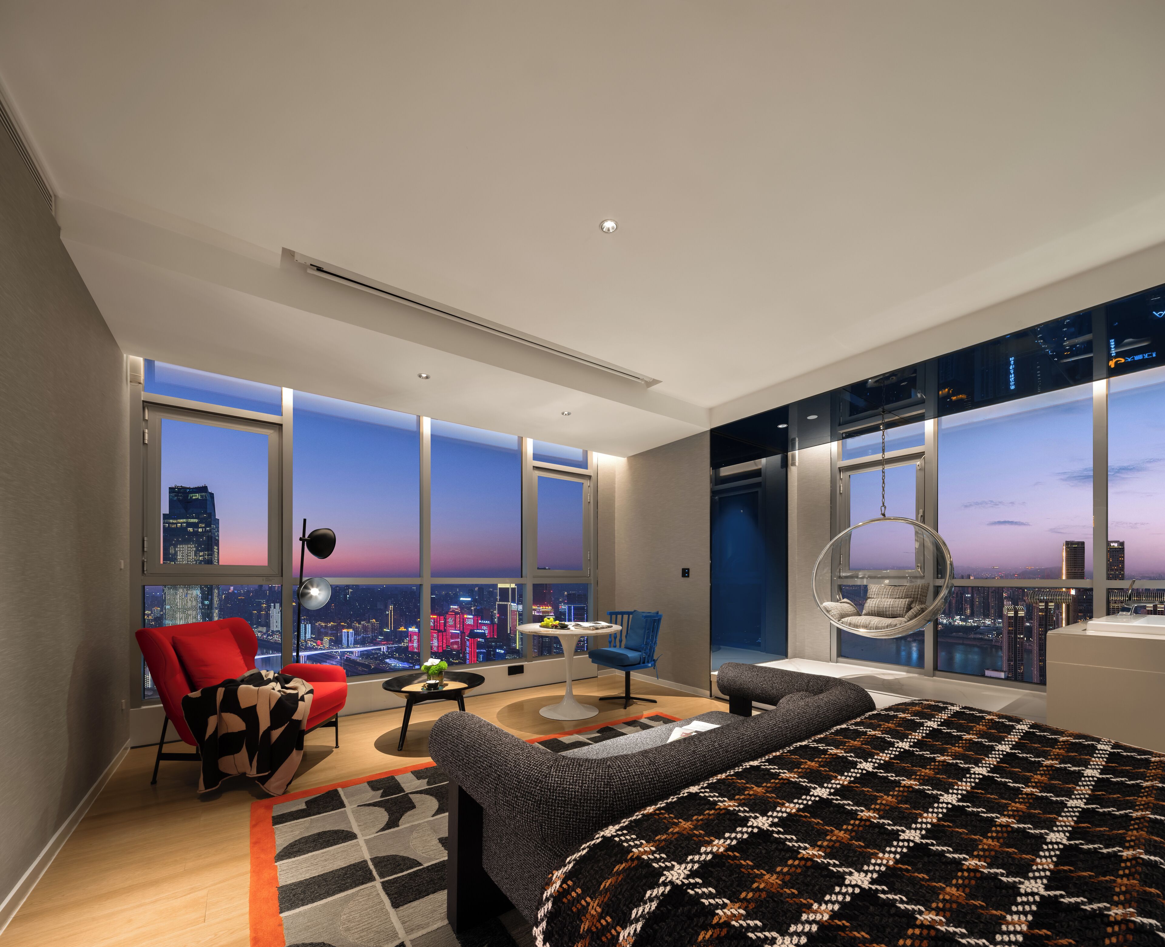 A modern hotel room with a large bed, a red chair, a blue chair, a round table, and a hanging chair.