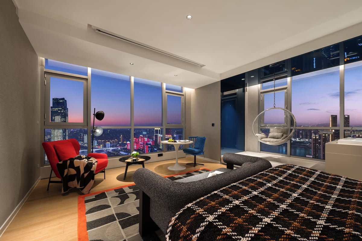 Una habitación de hotel moderna con una cama grande, una silla roja, una silla azul, una mesa redonda y una silla colgante.