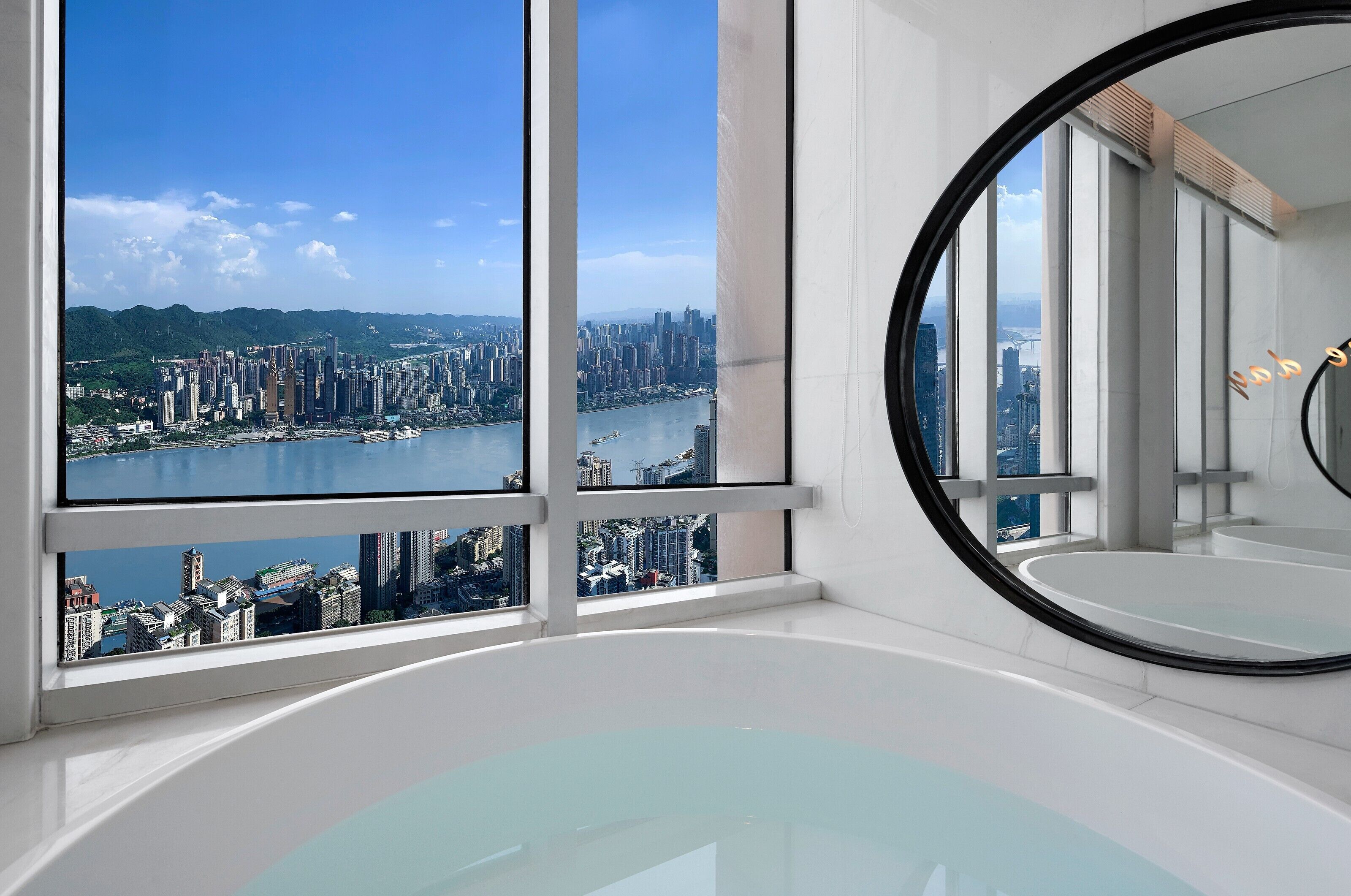 A modern hotel room with a large window offering a city view, a round mirror, and a bathtub.