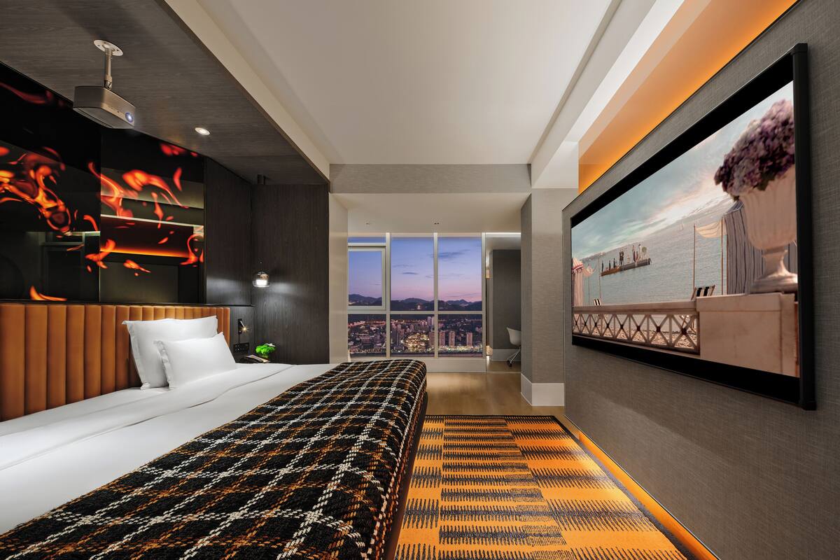 Une chambre d’hôtel moderne dotée d’un grand lit, offrant une vue sur la ville, d’un téléviseur à écran plat et d’une œuvre d’art murale originale.
