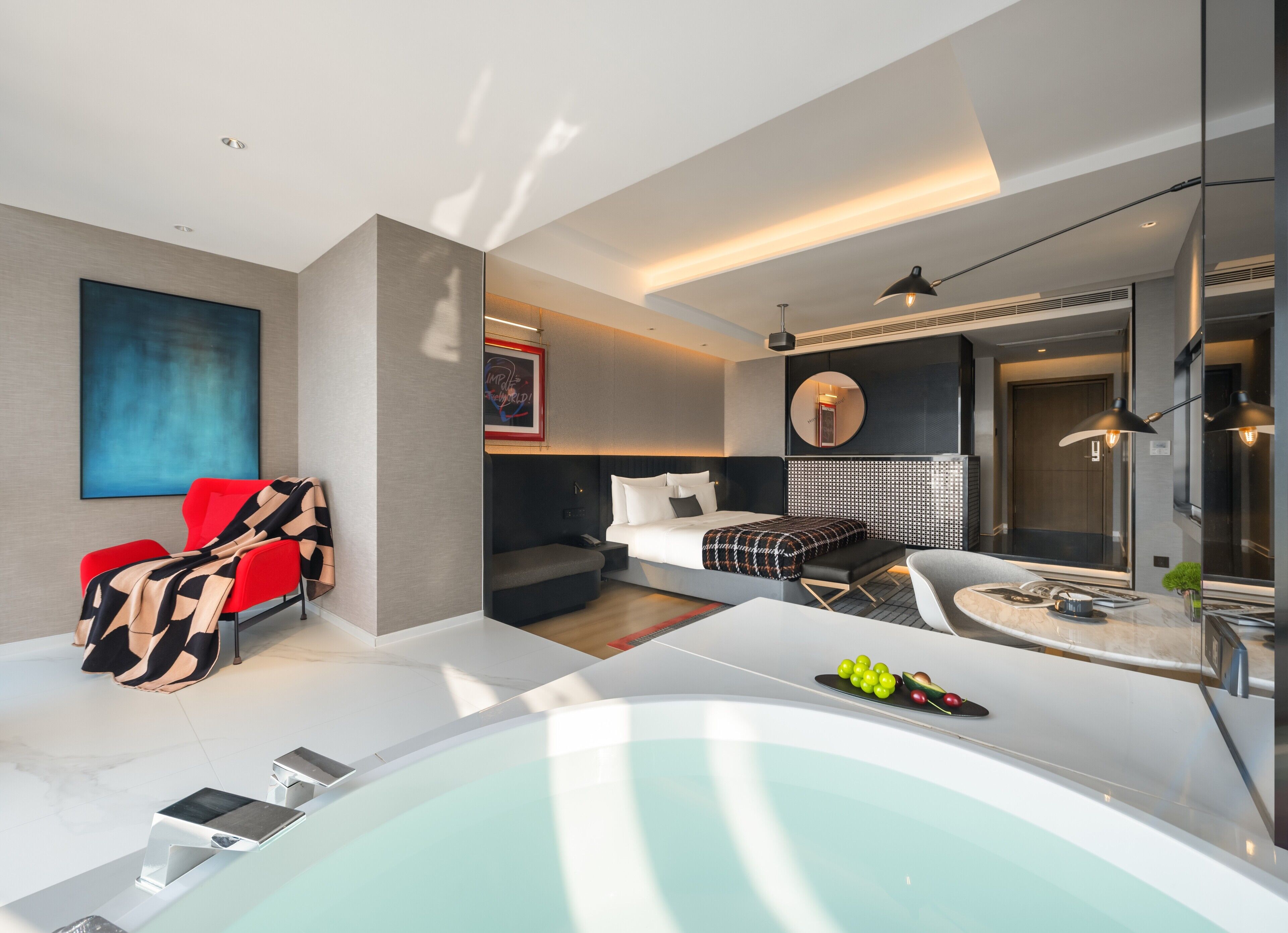 A modern hotel room with a large bathtub, a sofa, a red chair, a bed with a patterned blanket, and a wall-mounted TV.