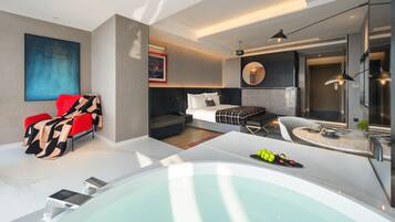 A modern hotel room with a large bathtub, a sofa, a red chair, a bed with a patterned blanket, and a wall-mounted TV.