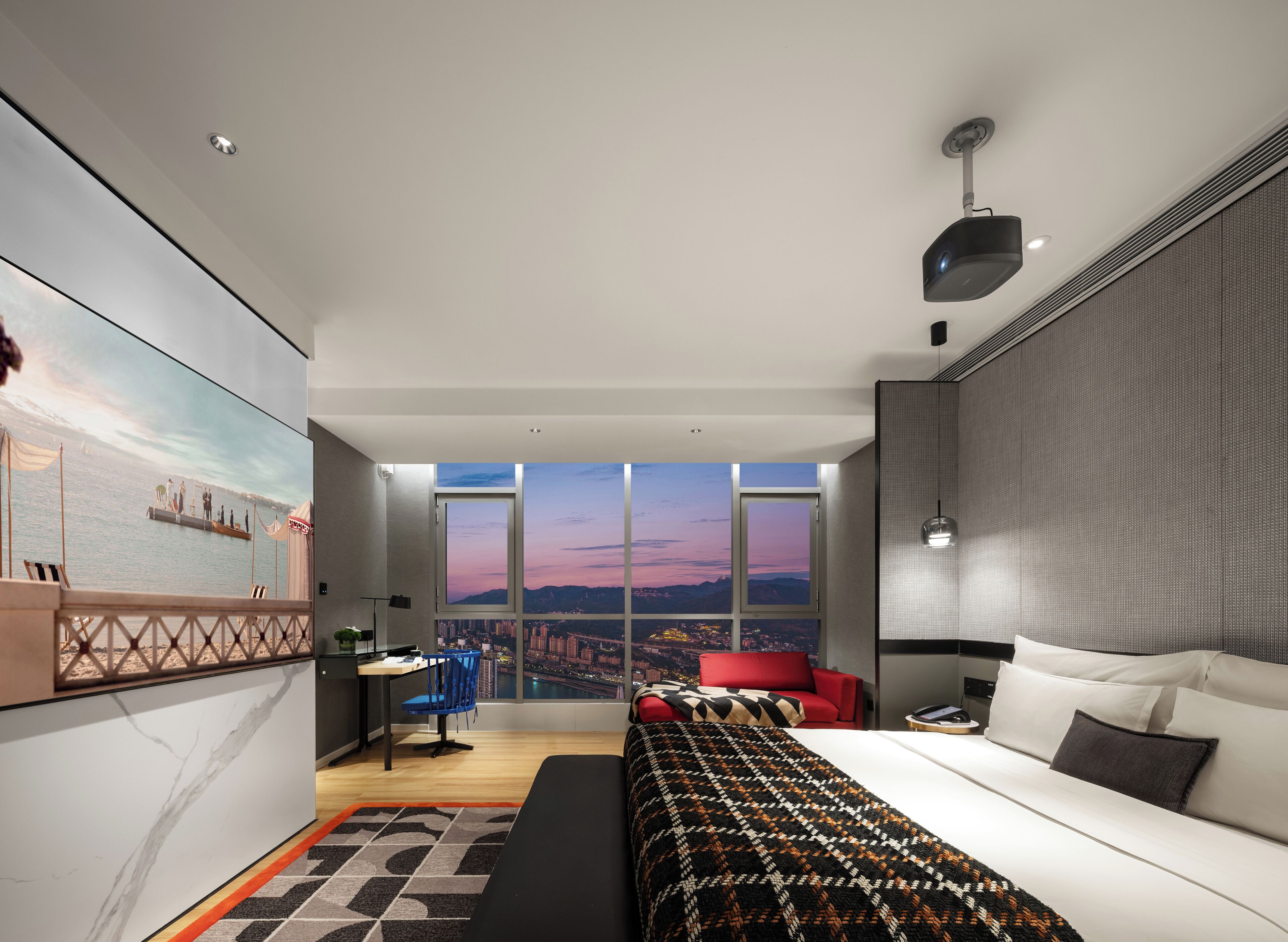 Una camera d'albergo moderna con un letto grande, una scrivania, una sedia rossa e vista sulla città attraverso ampie finestre.