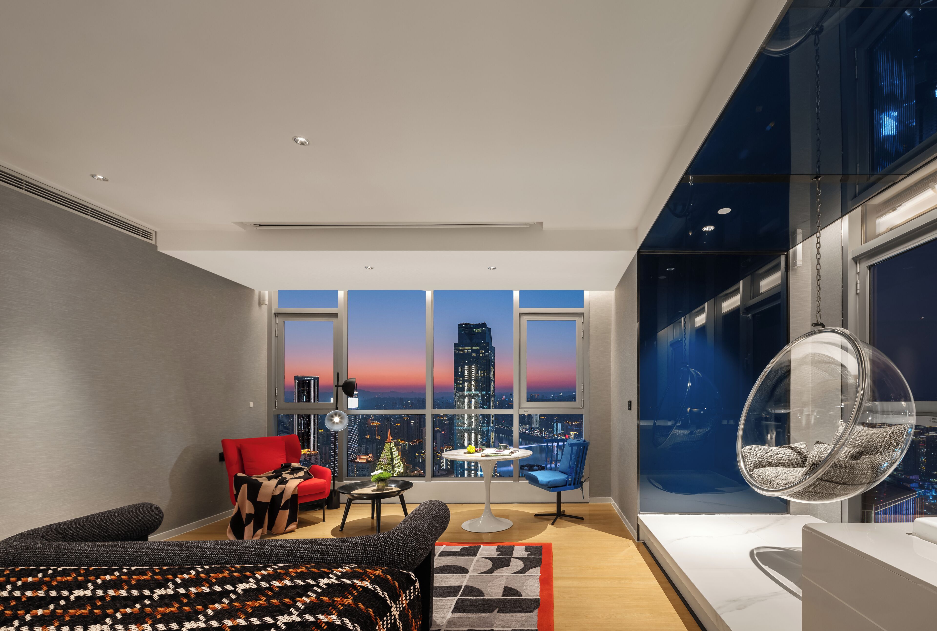 A modern room with a city view, a red chair, a hanging chair, and a small table with a lamp.