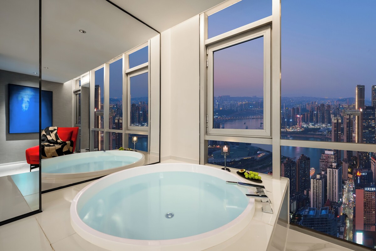 A modern bathroom with a large bathtub, a red chair, and a city view through large windows.