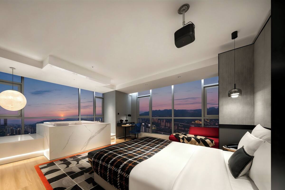 Une chambre moderne avec un grand lit, un bureau, une chaise, une grande fenêtre donnant sur la ville et un ciel illuminé par la lune.