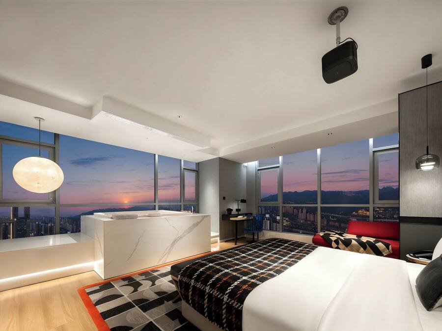 A modern bedroom with a large bed, a desk, a chair, a large window with a city view, and a moonlit sky.